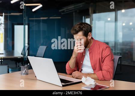 L'uomo malato al lavoro ha influenza e freddo, l'uomo d'affari starnutisce e tosse sul posto di lavoro che lavora all'interno dell'ufficio alla scrivania utilizzando un computer portatile al lavoro. Foto Stock