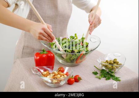 primo piano di insalata impastare con cucchiai di legno pomodori ciliegia cocktail cetrioli olive e altri ingredienti si trovano sul tavolo con tovaglia beige grembiule ristorante cibo che serve tavolo impostazione Foto Stock