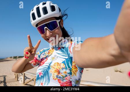 Ciclista felice in casco mostrando un gesto di pace e prendendo selfie Foto Stock