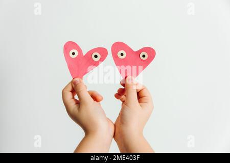 Fatto a mano per San Valentino o la giornata delle madri. Due cuori di carta rosa dipinti a mano con occhi nelle mani del bambino. Sfondo bianco. Foto Stock