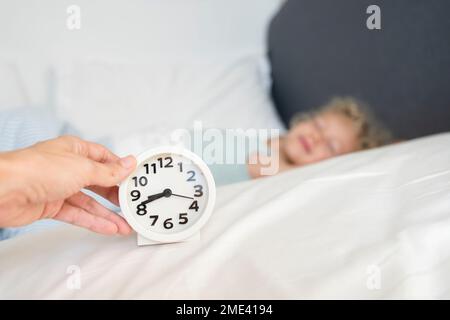 Mano di donna che tiene la sveglia di fronte a figlia che dorme sul letto Foto Stock