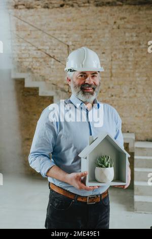 Felice uomo d'affari che tiene modello casa con vaso pianta in cantiere Foto Stock