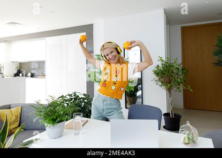 Donna allegra che indossa cuffie wireless che ballano con arance a casa Foto Stock