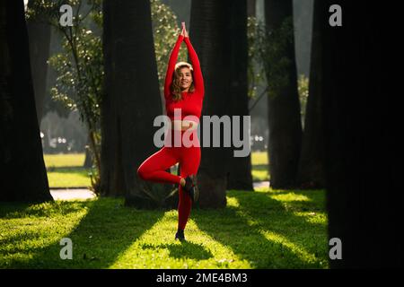 Donna con abito yoga rosso, posa albero yoga su sfondo verde. Foto Stock