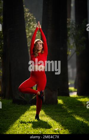 Donna con abito yoga rosso, posa albero yoga su sfondo verde. Foto Stock