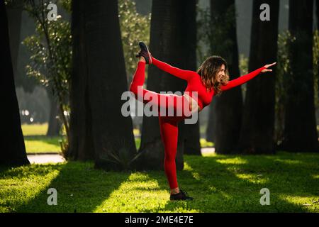 Donna con abito yoga rosso, posa yoga Natarajasana signore della danza su sfondo verde grasss. Foto Stock