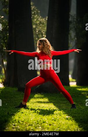 Donna con abito da yoga di colore rosso, che posa movimenti di yoga su uno sfondo verde grasss. Foto Stock