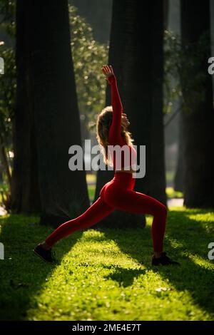 Donna con abito da yoga di colore rosso, che posa i movimenti del guerriero di yoga su uno sfondo verde grasss Foto Stock