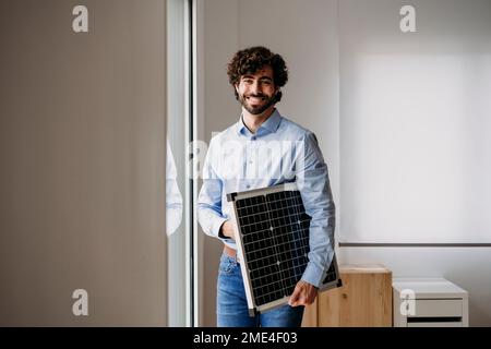 Felice uomo d'affari giovane in piedi con pannelli solari vicino alla finestra in ufficio Foto Stock