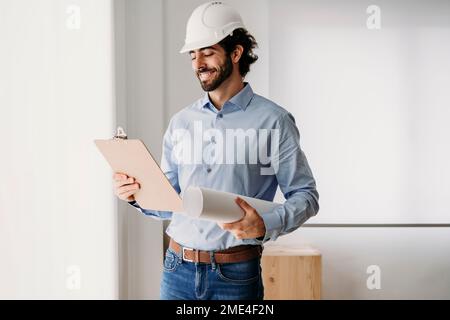 Architetto sorridente che tiene i progetti dalla finestra in ufficio Foto Stock