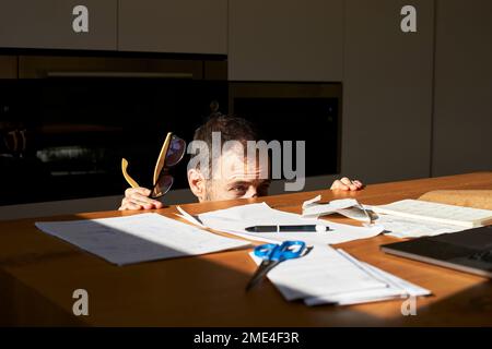 Uomo preoccupato che si nasconde dalle fatture finanziarie sulla scrivania a casa Foto Stock