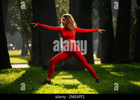 Donna con abito da yoga di colore rosso, che posa movimenti di yoga su uno sfondo verde grasss. Foto Stock