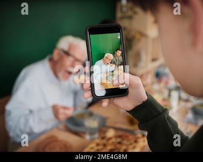 Nipote filma padre e nonno con un telefono mentre pranzano Foto Stock