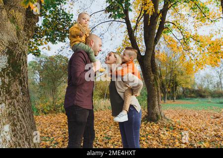 Donna sorridente che dà cavalcata piggyback a figlio e uomo che porta il ragazzo sulle spalle nella foresta autunnale Foto Stock