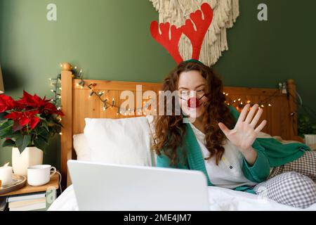 Donna che indossa corna e naso di clown che sventola durante la videochiamata attraverso il computer portatile in camera da letto Foto Stock