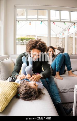 Felice padre solleticare la figlia sul divano con la madre seduta in background Foto Stock
