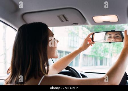 Giovane donna che regola lo specchietto retrovisore in auto Foto Stock