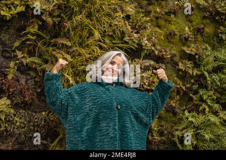 Donna anziana felice che flette i muscoli di fronte alle piante Foto Stock