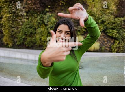 Donna sorridente che fa cornice con le dita davanti al giardino verticale Foto Stock