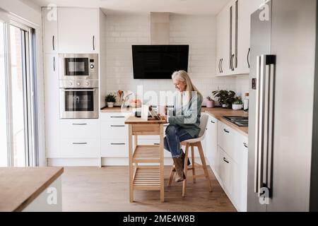 Donna matura che utilizza un tablet PC seduto in cucina a casa Foto Stock