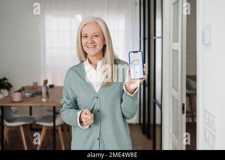 Donna felice che mostra l'app mobile del termostato sullo schermo del dispositivo a casa Foto Stock