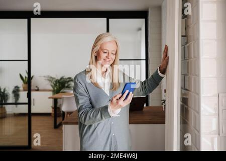 Donna sorridente che usa il telefono cellulare e tocca il radiatore a casa Foto Stock