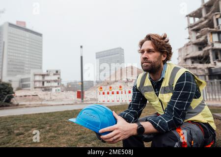 Lavoratore maturo contemplativo seduto con elmetto in cantiere Foto Stock