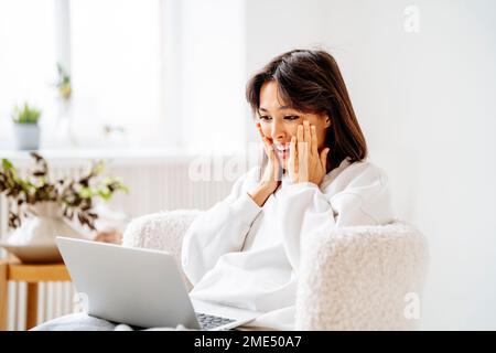 Sorpresa giovane donna che guarda il notebook seduto sulla sedia Foto Stock