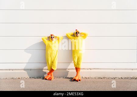 Donna e uomo che indossano costumi di pollo appoggiati con le mani dietro la testa sul muro Foto Stock