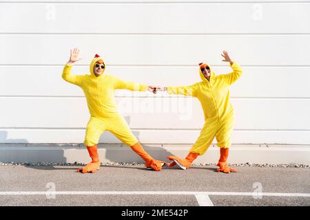 Amici giocosi che indossano costumi di pollo in mano e ballano davanti al muro Foto Stock