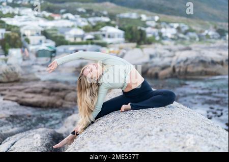 Giovane donna sorridente che fa esercizi di stretching sulla roccia dal mare Foto Stock