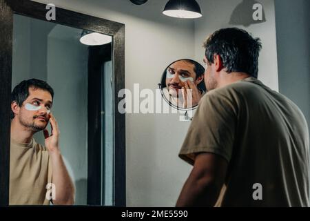 Uomo che applica le zone sotto gli occhi davanti allo specchio Foto Stock