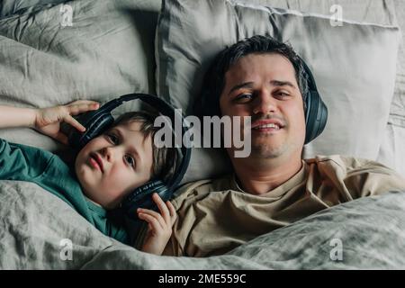 Padre e figlio indossano cuffie che ascoltano la musica a letto Foto Stock