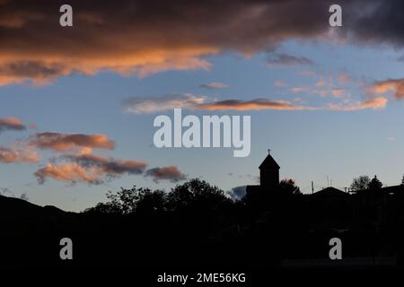 Silhouette di campanile della chiesa cattolica che si staglia sopra le cime degli alberi con il tramonto nuvole arancioni nel cielo nella città di Akhaltsikhe, Georgia. Foto Stock