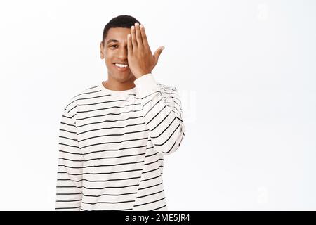 Un uomo afroamericano sorridente copre metà del volto, controlla gli occhi presso il negozio ottico, si erge su sfondo bianco Foto Stock