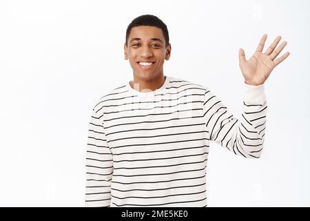 Sorridente afroamericano che saluta la mano, agita con un volto amichevole e felice, sfondo bianco Foto Stock