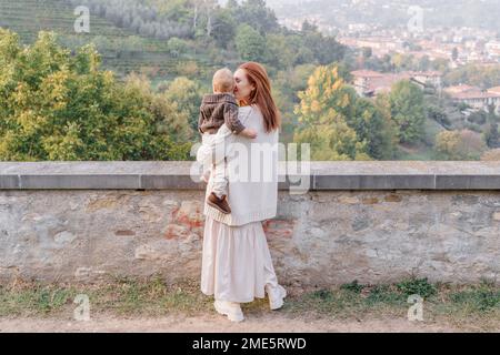 Una giovane donna con i capelli rossi tiene il figlio del bambino fra le braccia e lo abbraccia teneramente. Si levano in piedi con le loro spalle contro lo sfondo del hil verde Foto Stock