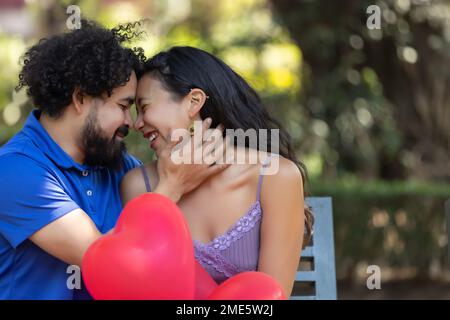 giovane coppia messicana latina ridendo e parlando il giorno di san valentino, con palloncini a forma di cuore rosso Foto Stock