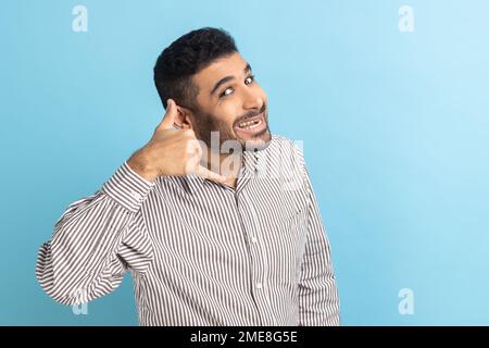 Ci contatti. Ritratto di uomo d'affari positivo con barba facendo gesto con le dita comporre il mio numero o richiamare me, indossando camicia a strisce. Studio in interni isolato su sfondo blu. Foto Stock