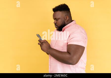 Vista laterale di un serio uomo che indossa una camicia rosa inviando messaggi di testo tramite telefono cellulare, cerca regali per le vacanze in Internet, utilizza l'app per smartphone. Studio al coperto isolato su sfondo giallo. Foto Stock