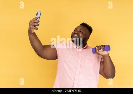 Ritratto di giovane adulto allegro soddisfatto in camicia rosa in piedi con manubri e fare selfie mentre si fa esercizio, la trasmissione in diretta. Studio al coperto isolato su sfondo giallo. Foto Stock