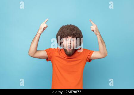 WOW, sguardo, pubblicità pazzesca. Uomo con acconciatura Afro in T-shirt arancione che punta in su a posto vuoto per il contenuto dell'annuncio ed esprimere lo stupore. Studio in interni isolato su sfondo blu. Foto Stock