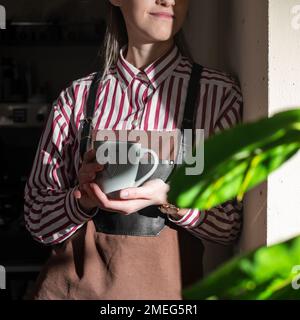 La giovane barista ragazza vicino a una moderna finestra gode di una tazza di caffè, guardando fuori, Buongiorno, giornata di lavoro, libero dal lavoro e dai doveri. Foto Stock