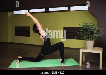 Attraente donna di mezza età, che conduce uno stile di vita sano e pratica yoga, esegue l'esercizio Anjaneyasana, una posa crescente, si allena da sola nella st Foto Stock