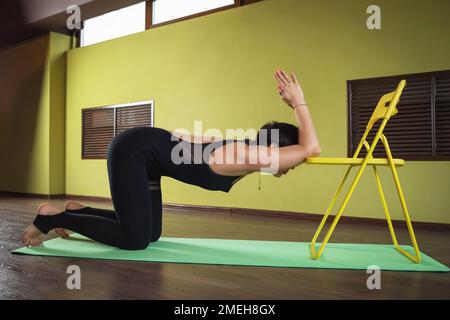 Attraente donna di mezza età che pratica yoga esegue vari esercizi per allungare i muscoli della colonna vertebrale e delle spalle con l'aiuto di una sedia Foto Stock