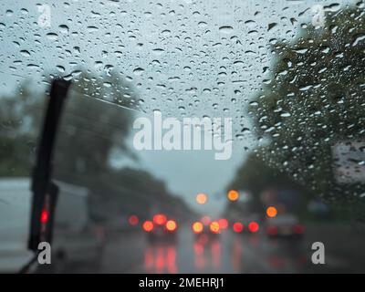 Primo piano delle gocce di pioggia su un parabrezza, tergicristalli sfocati che puliscono il vetro e traffico stradale con luci auto sfocate. Vista dal sedile di guida. Foto Stock
