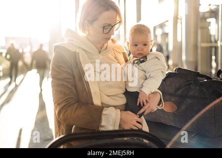 Motherat viaggia con il bambino, camminando, spingendo passeggino e carretto bagagli davanti alla stazione del terminal aeroportuale Foto Stock