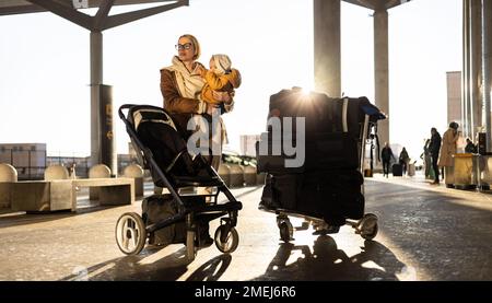 Motherat viaggia con il bambino, camminando, spingendo passeggino e carretto bagagli davanti alla stazione del terminal aeroportuale Foto Stock