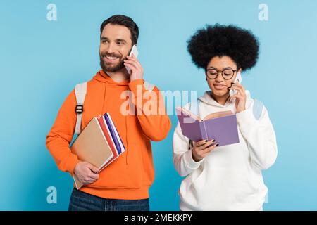 sorridenti studenti multietnici in cappuccio arancione e bianco che parlano su telefoni cellulari isolati su blu Foto Stock