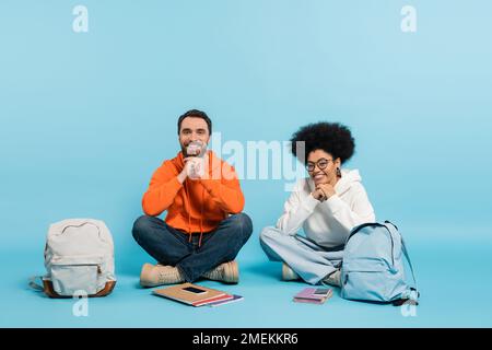 felici studenti multietnici seduti con gambe incrociate vicino a zaini e notebook con smartphone in blu Foto Stock